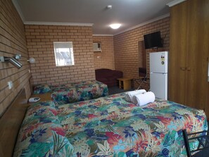 A room with a brick wall, a bed with floral bedding, a red sofa, a small table, a TV mounted on the wall, and a refrigerator.