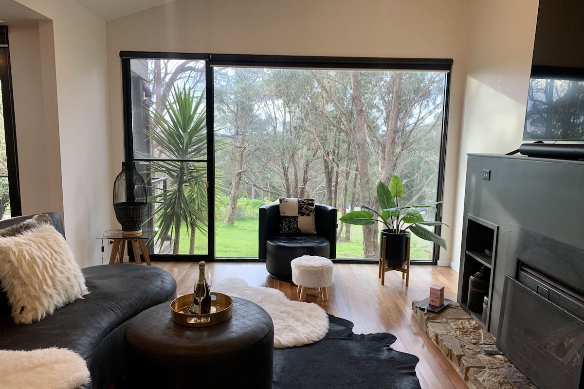 A modern living room with a black sofa, a round ottoman, a fireplace, and a large sliding glass door leading to a garden.