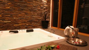 Zona de spa con pared de piedra, bañera, pétalos de rosa, velas y una bandeja con vaso de agua.
