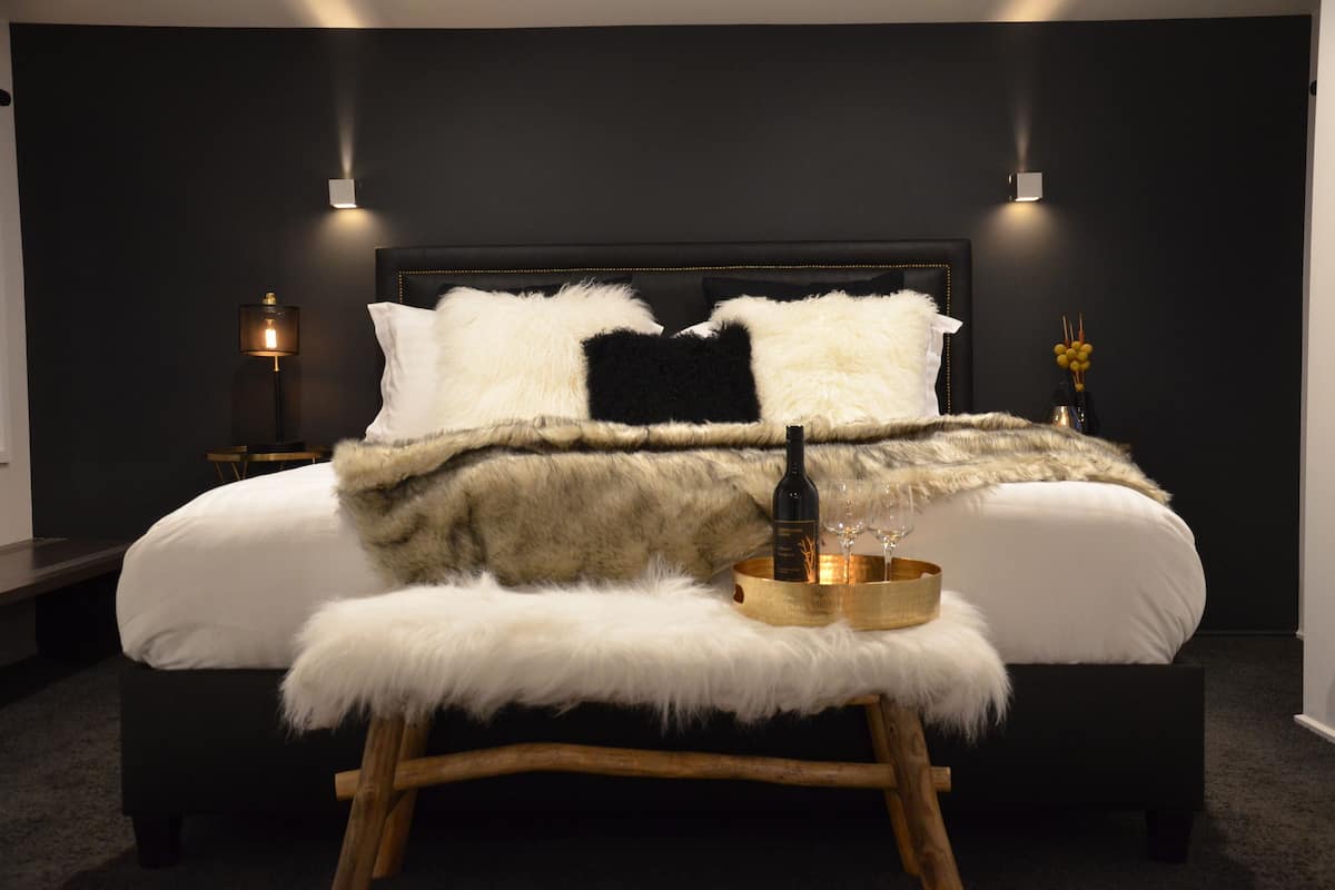 A modern bedroom with a dark headboard, white bedding, fluffy pillows, a bottle of wine, and a candle holder on a wooden stool.
