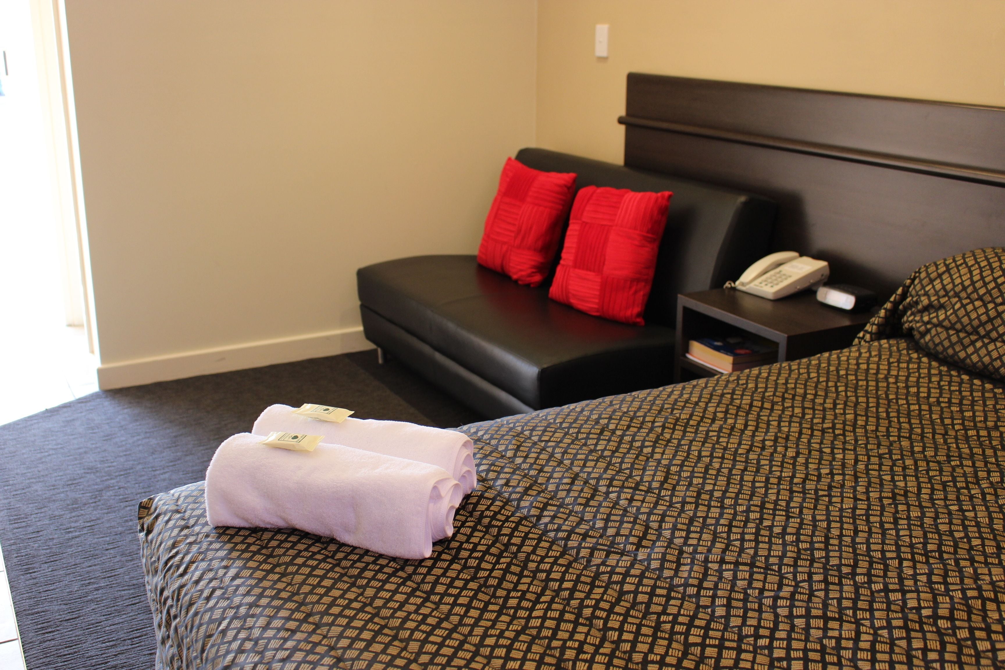 A hotel room with a bed, a small sofa, a nightstand with a phone, and red pillows.