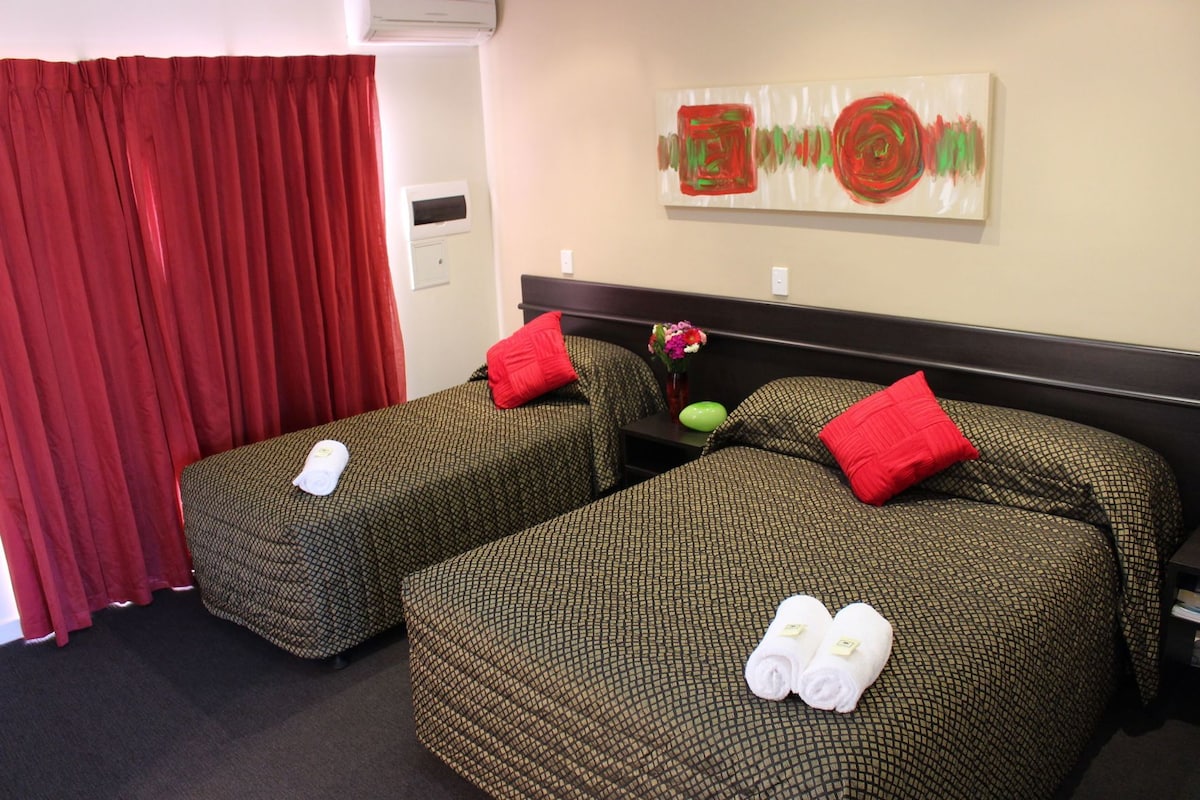 Habitación de hotel con dos camas, cortinas rojas, una obra de arte en la pared y una mesita con un jarrón de flores.
