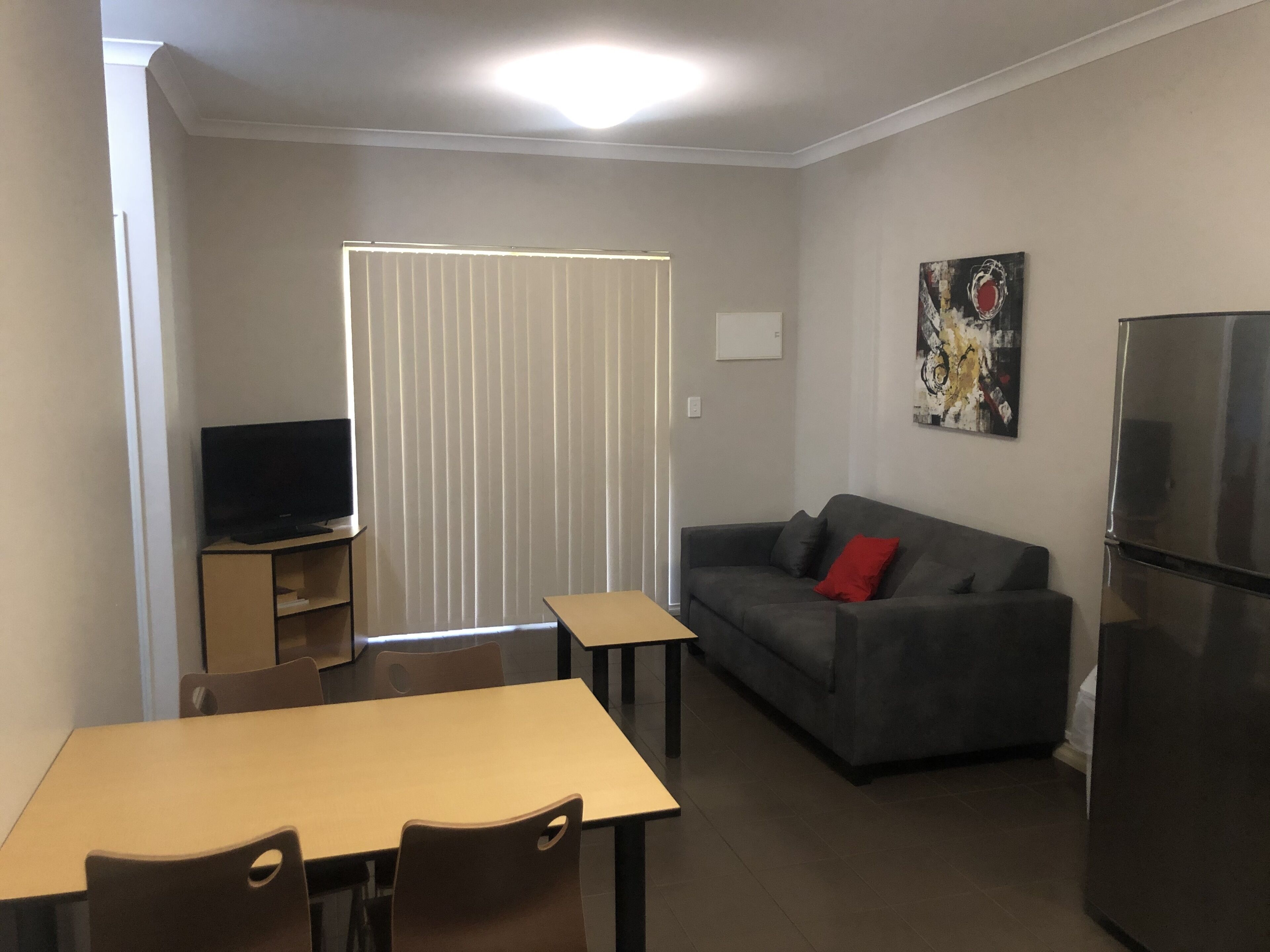 A compact living space with a dining table, two chairs, a sofa with a red pillow, a television on a stand, and a refrigerator.