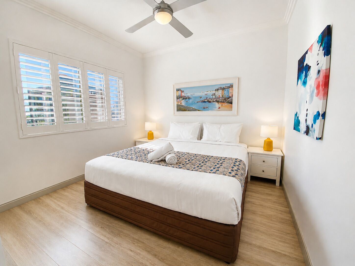 A bedroom with a bed, two bedside tables, a ceiling fan, a window with blinds, and a painting on the wall.