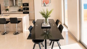 A modern kitchen with stainless steel appliances, a dining area with a glass table and black chairs, and a large window offering a view of the sea. - Redvue Apartments (Redcliffe)