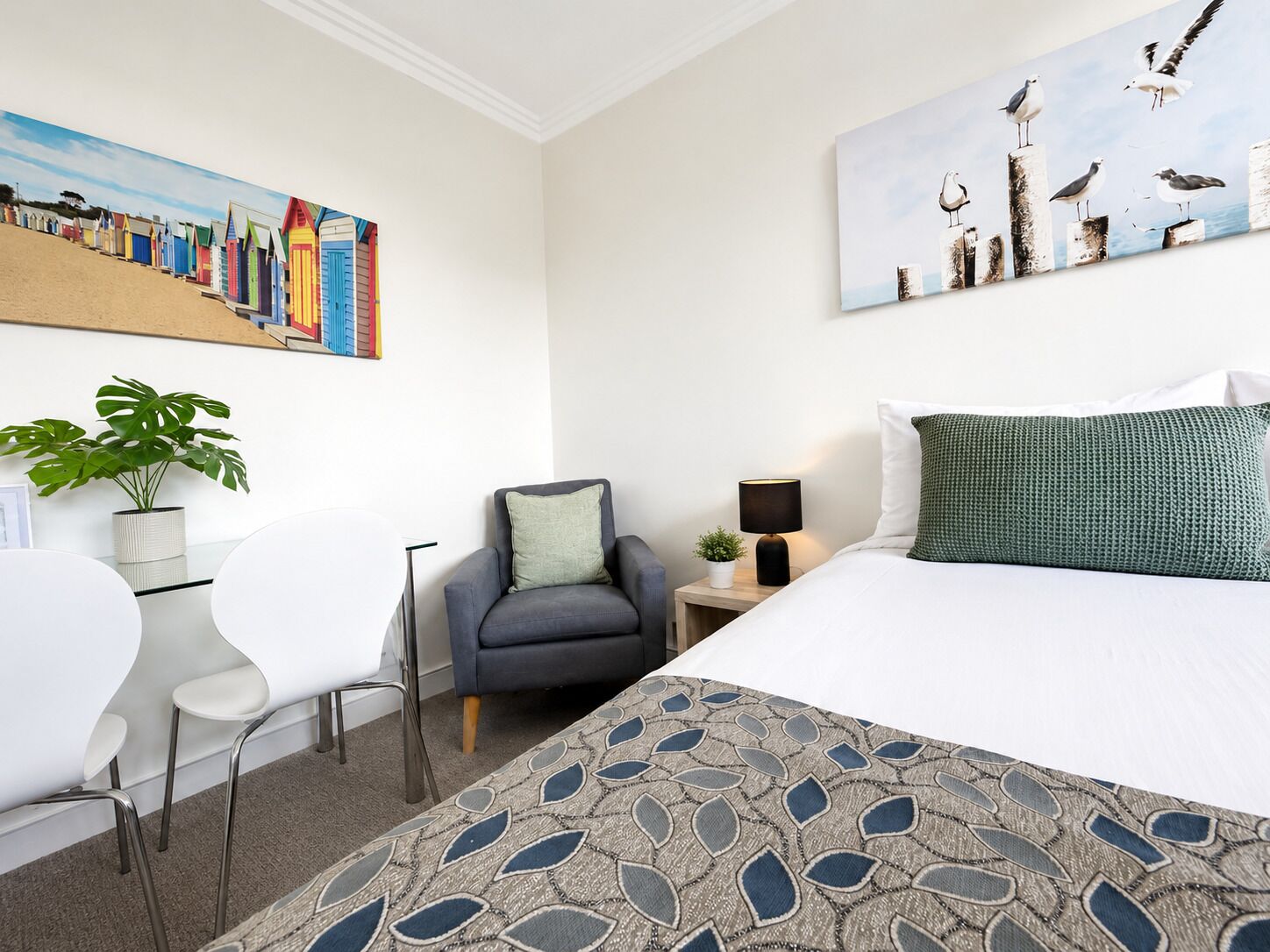 Una habitación de hotel con cama, escritorio, silla, planta en maceta y dos cuadros en la pared.