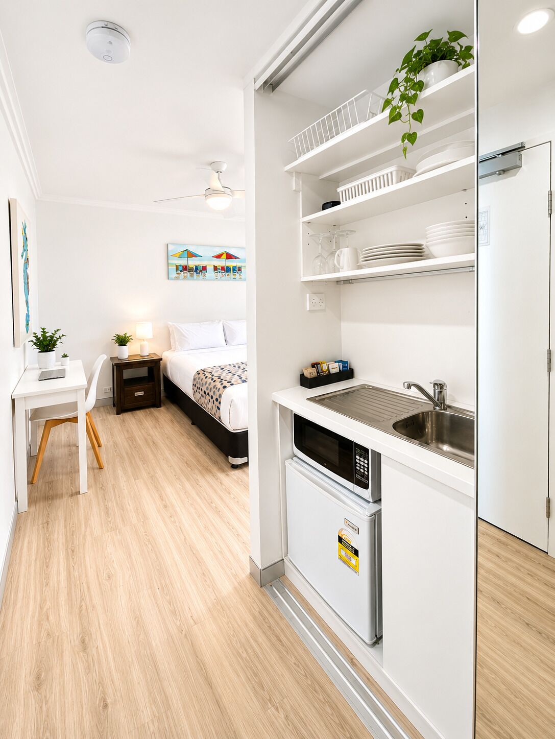Una habitación de hotel compacta con una cama, mesita de noche, lámpara y un escritorio pequeño con una silla. Cuenta con una kitchenette con un lavamanos, microondas y armarios de almacenamiento.