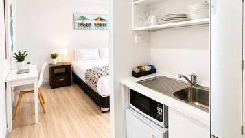 Una habitación de hotel compacta con una cama, mesita de noche, lámpara y un escritorio pequeño con una silla. Cuenta con una kitchenette con un lavamanos, microondas y armarios de almacenamiento.