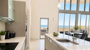 A modern kitchen with a large island, a view of the sea, and a dining area with outdoor seating. - Redvue Apartments (Redcliffe)