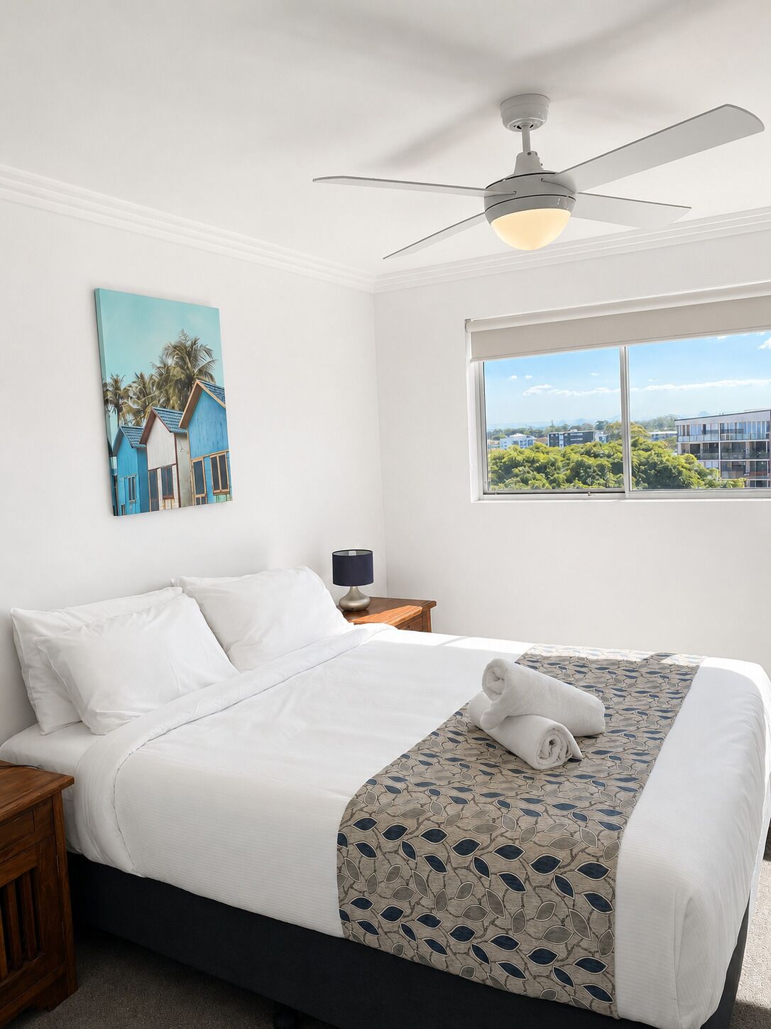 Habitación de hotel con cama, ventilador de techo, ventana con vistas y un cuadro en la pared.