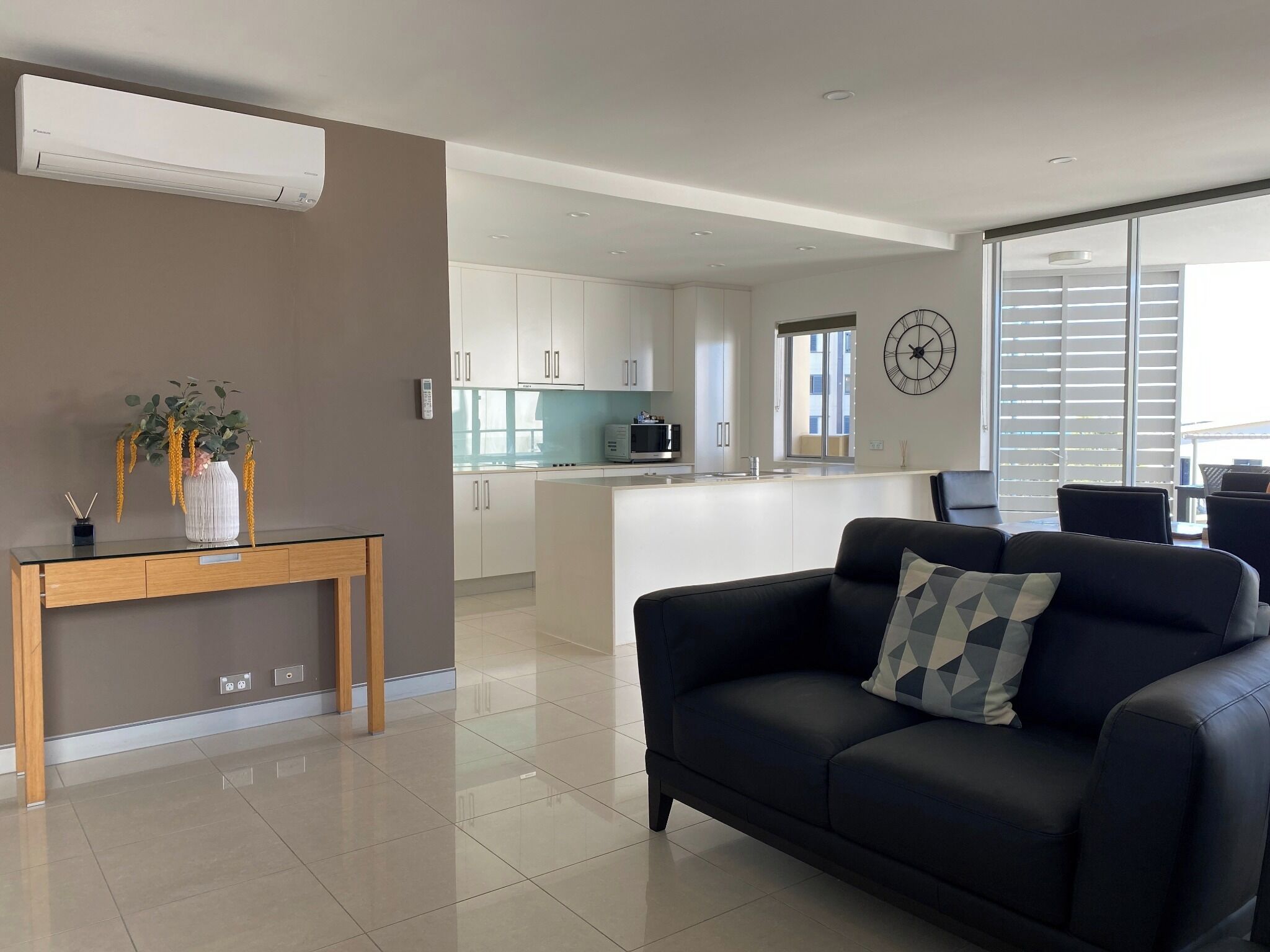 Una sala moderna con un sofá de cuero negro, una mesita de madera con un florero, y una cocina al fondo con armarios blancos y un ventanal grande.