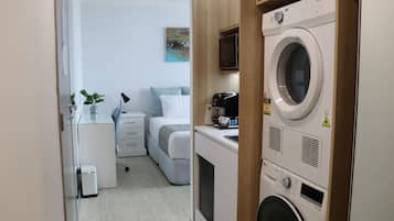 Habitación de hotel compacta con lavadora, cama con cabecero gris, mesita de noche con lámpara y pequeño escritorio con planta.