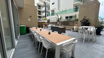 Una terrazza esterna con un lungo tavolo di legno e sedie bianche, circondata da piante in vaso e un angolo cottura.