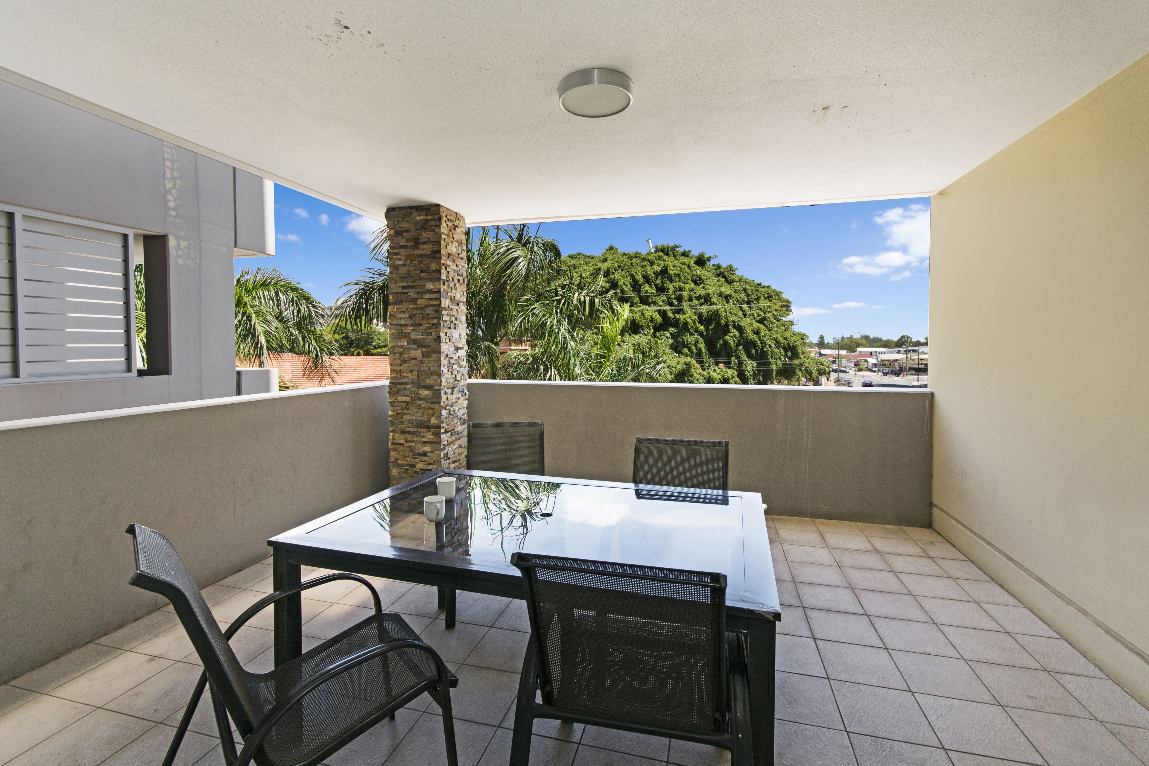A balcony with a glass-top table and chairs, a stone pillar, and a view of a green hill and buildings.