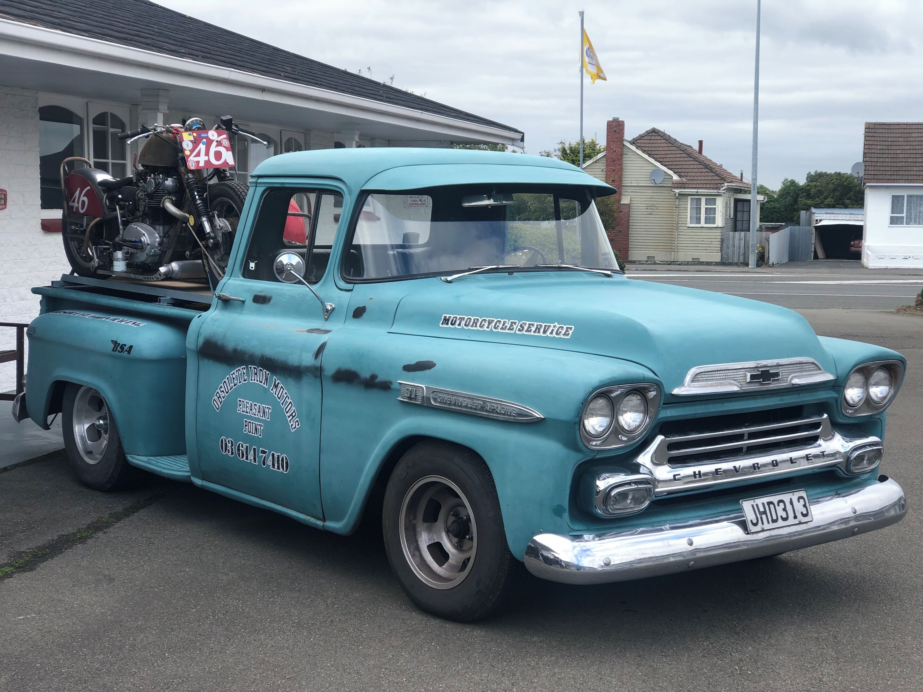 A teal vintage Chevrolet truck with a motorcycle mounted on the back, parked in a residential area.