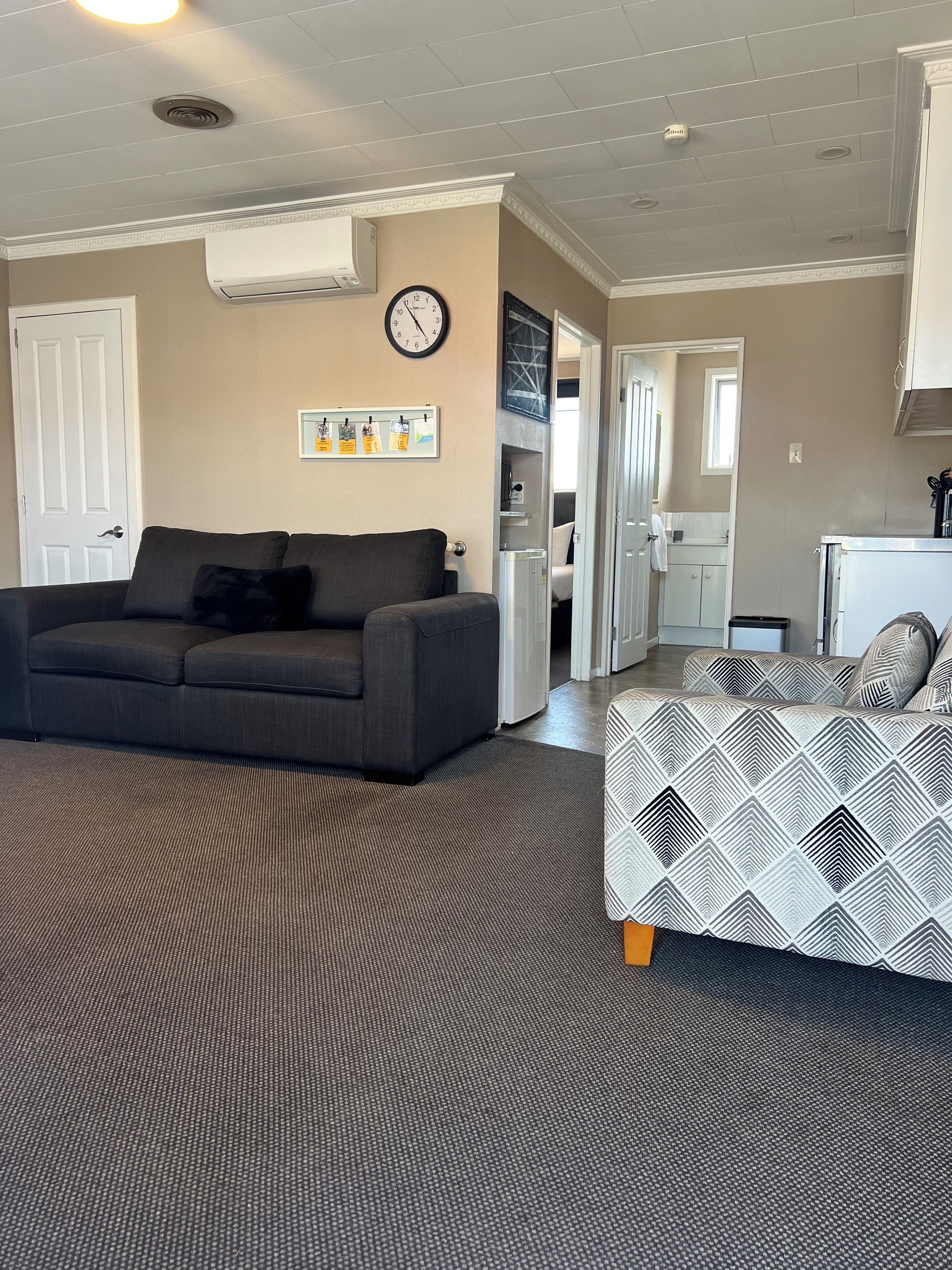 A living room with a grey sofa, a patterned sofa, a clock, and a wall-mounted air conditioner.