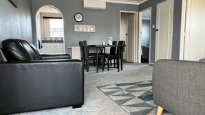 Una sala de estar con un sofá de cuero negro, un sofá gris y un comedor con una mesa y sillas. Hay un reloj en la pared y un radiador sobre la mesa.