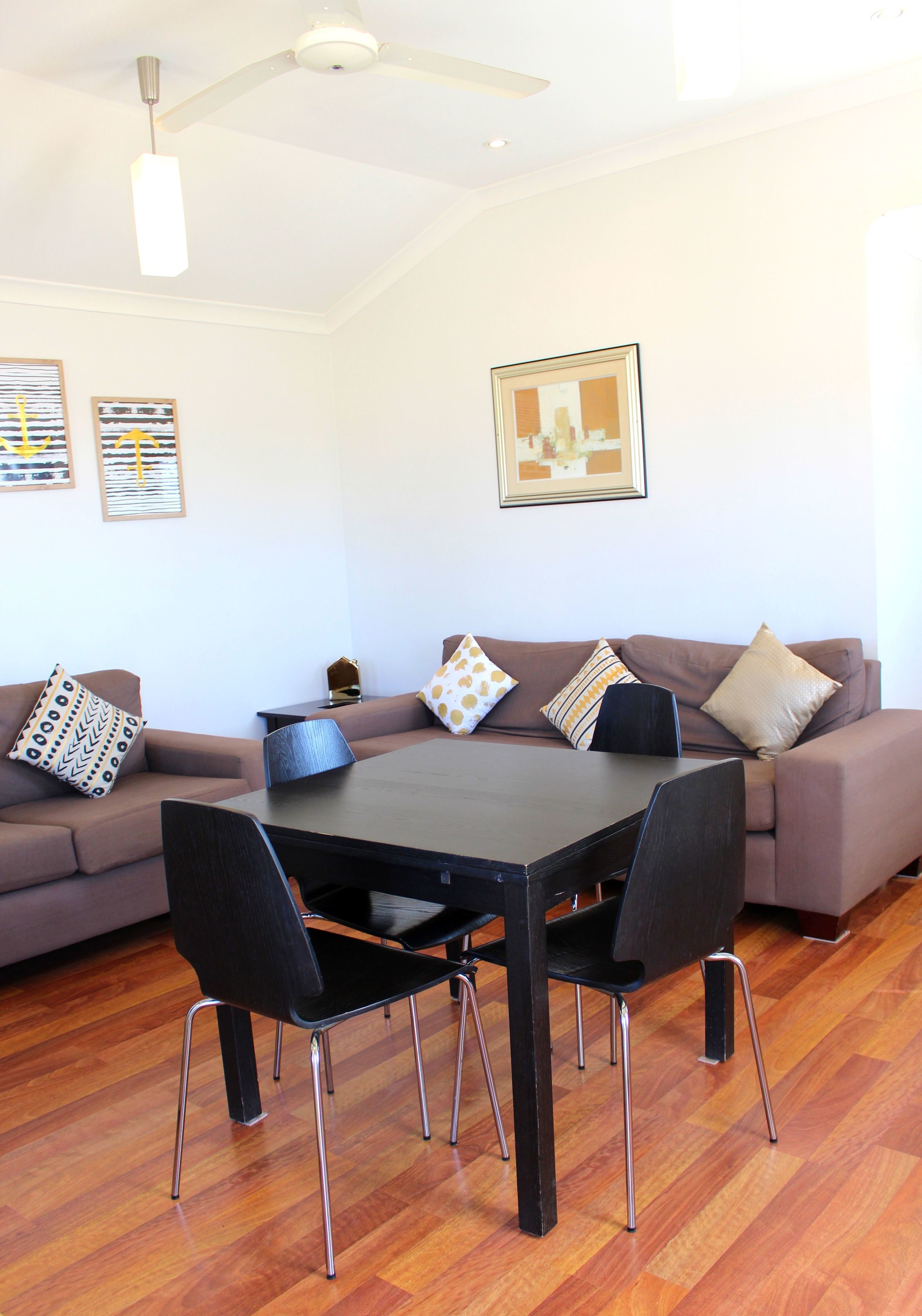 A modern living room with a dark wooden dining table, black chairs, a brown sofa, and a ceiling fan.