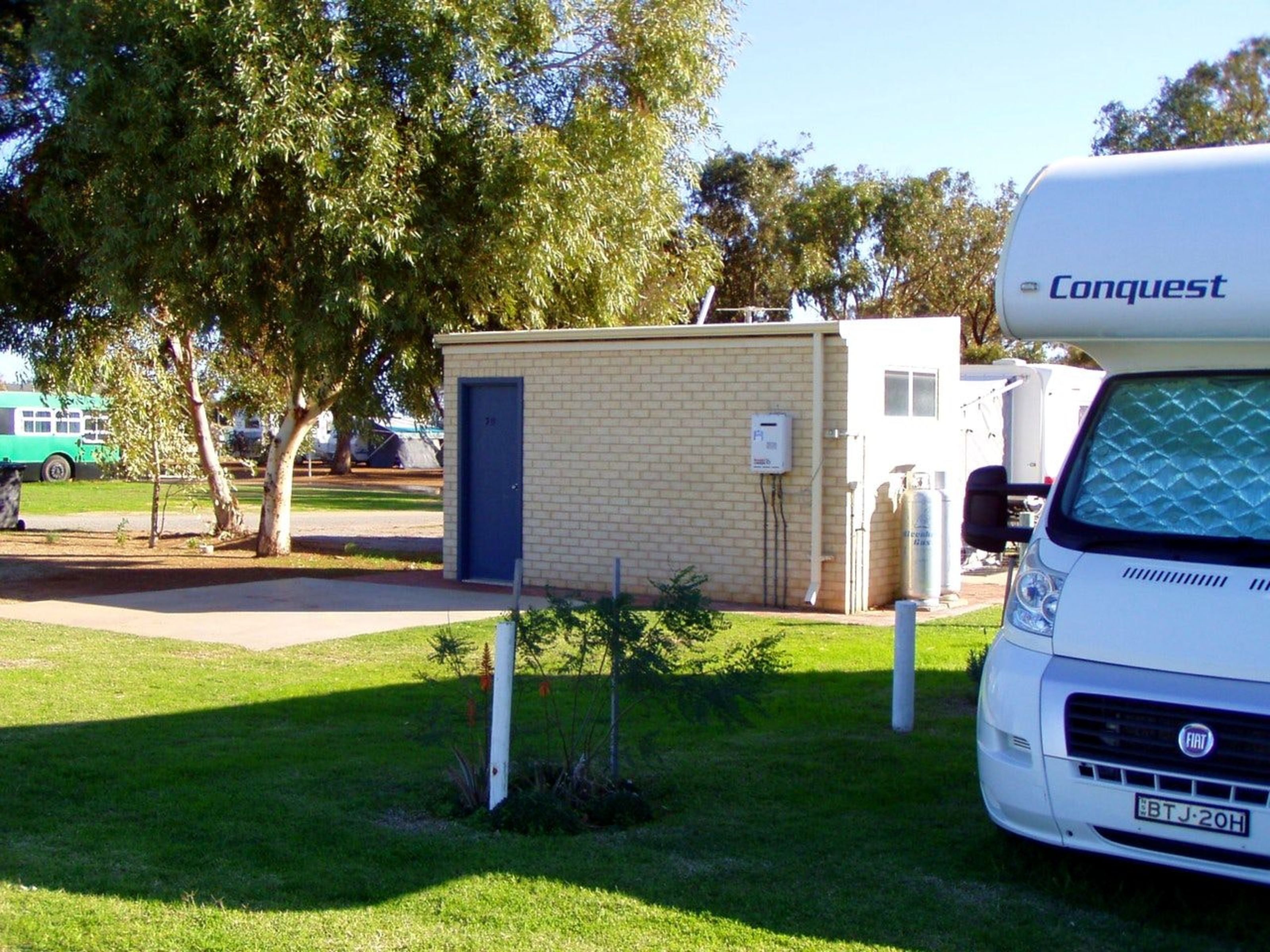 A white RV parked in a grassy area with a brick building labeled 'Conquest' in the background.
