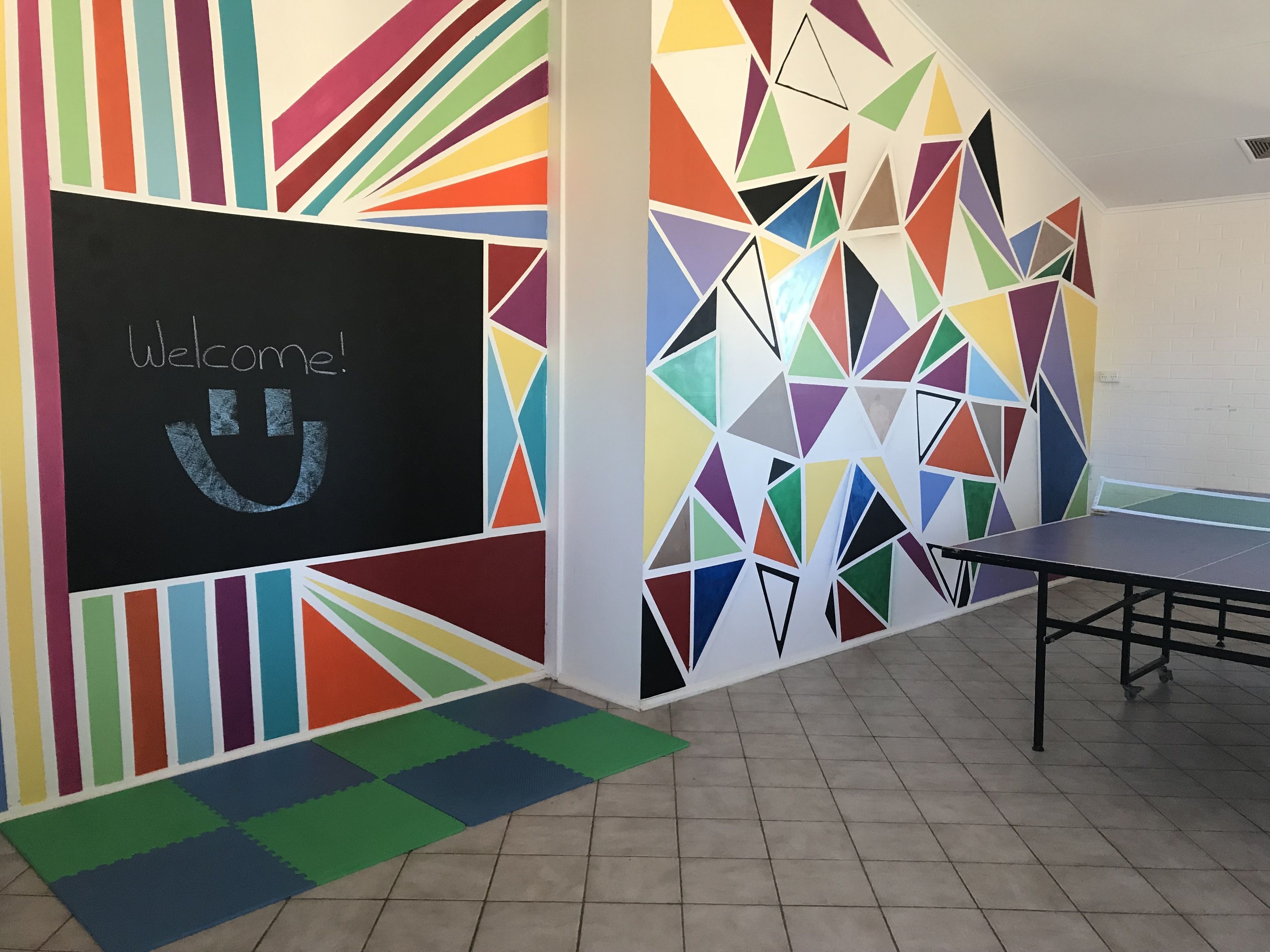 A room with a colorful geometric wall design, a welcome sign, and a ping pong table.