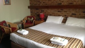 A hotel room with a large bed, a brick wall, a wicker chair, and a painting on the wall. - Gisborne Motel (Gisborne)