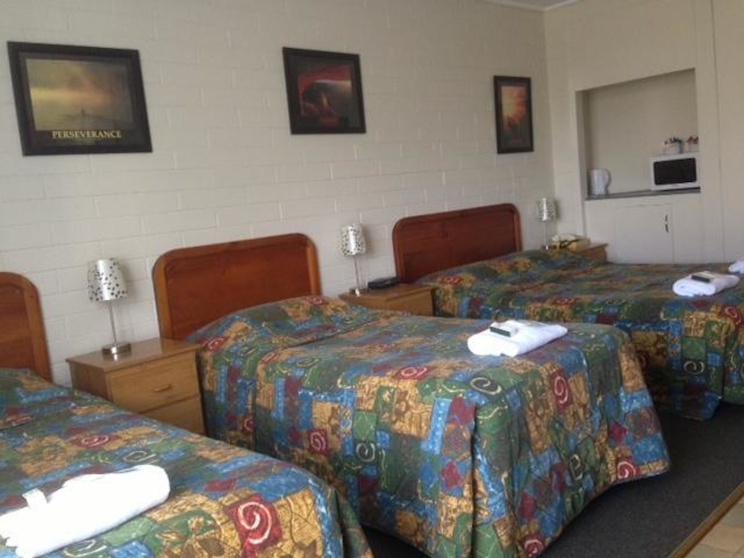 A hotel room with three beds, each with a colorful patterned bedspread. There are framed pictures on the wall, a small desk with a lamp, and a microwave above a cabinet.