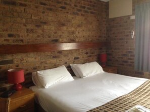 Une chambre d’hôtel avec un mur en briques, une tête de lit en bois, un grand lit avec une literie blanche, deux tables de chevet et une lampe rouge sur l’une des tables.