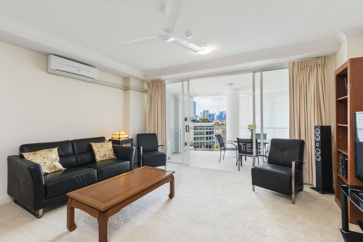 A modern living room with a black leather sofa, a wooden coffee table, and a black armchair. There is a flat-screen TV on a wooden stand and a ceiling fan. The room has a large sliding glass door leading to a balcony with a city view.