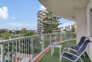 A balcony with two lounge chairs and a small table, overlooking a green artificial lawn and a cityscape with tall buildings. - Founda Gardens Apartments (Auchenflower)