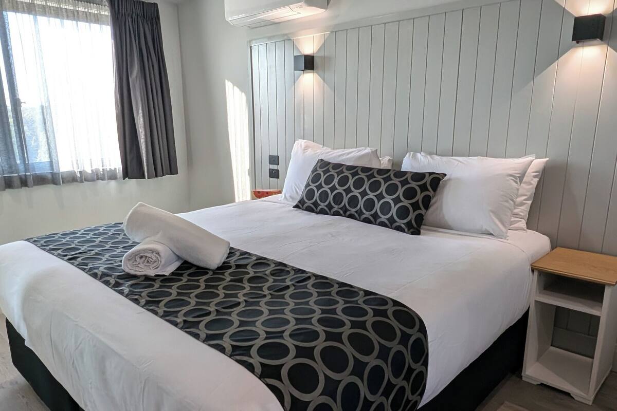 A modern hotel room with a large bed, a bedside table, an air conditioning unit, and a window with curtains.