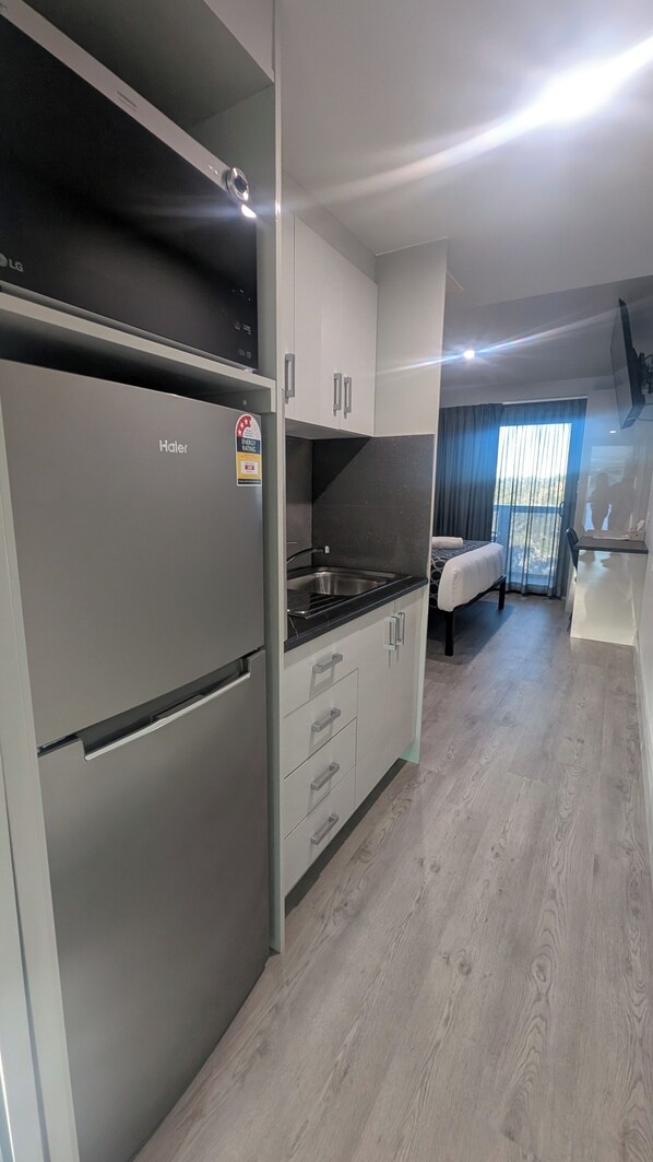 A modern kitchen with a built-in LG refrigerator, a stainless steel sink, and a black countertop. There is a bed with a window view in the adjacent room.