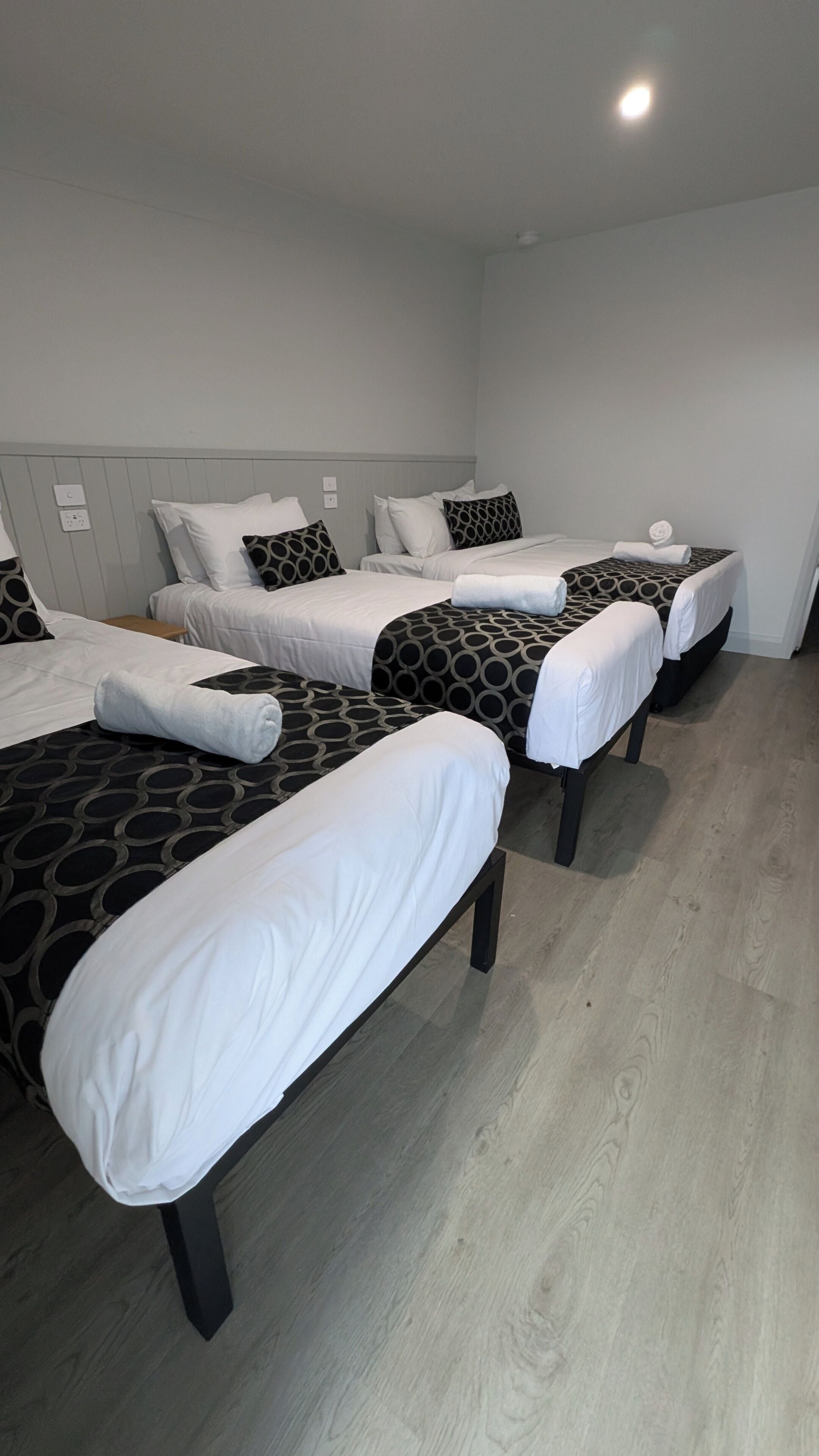 Habitación de hotel con tres camas individuales, cada una con un edredón estampado y almohadas blancas. El piso es de madera y el techo blanco tiene una lámpara.