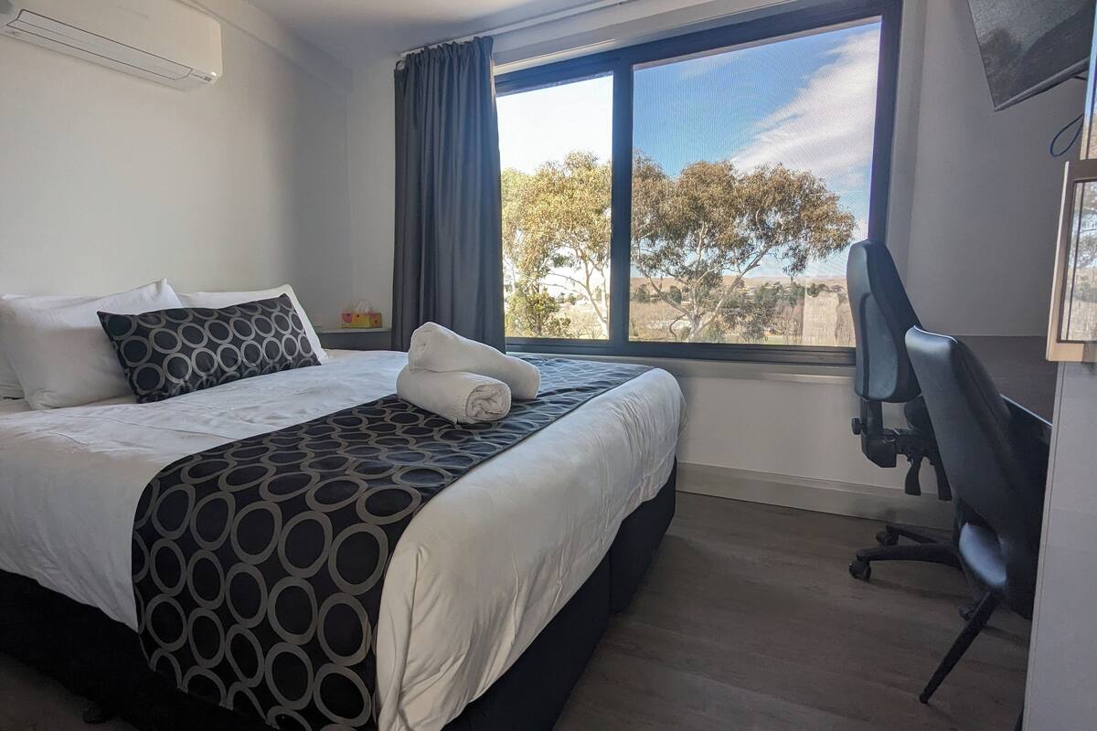 A neatly made bed with white and patterned linens, a window with a view of trees, and a desk with a chair.