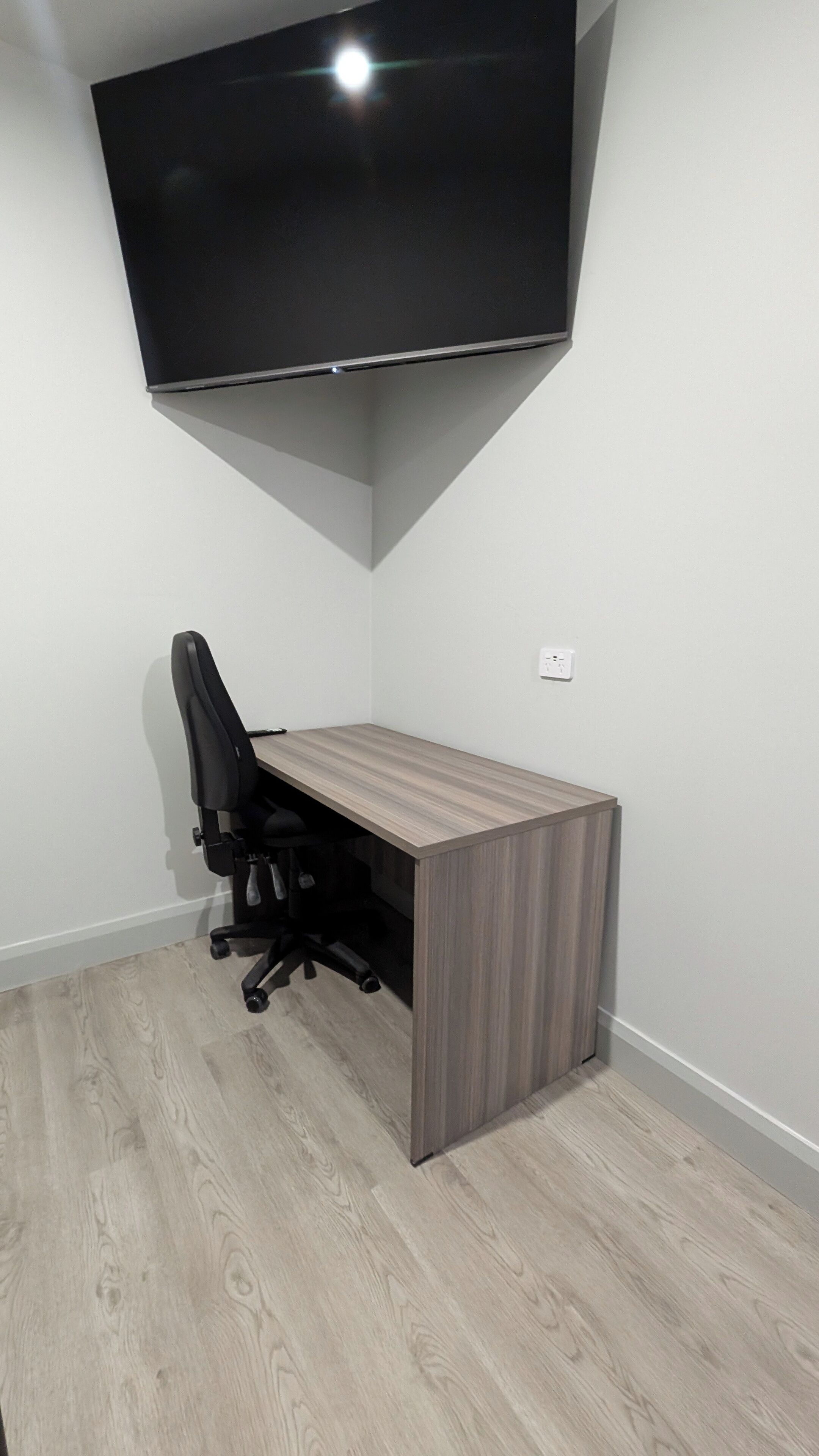 Un bureau d’angle avec un téléviseur à écran plat fixé au mur, une chaise de bureau noire et un bureau en bois.