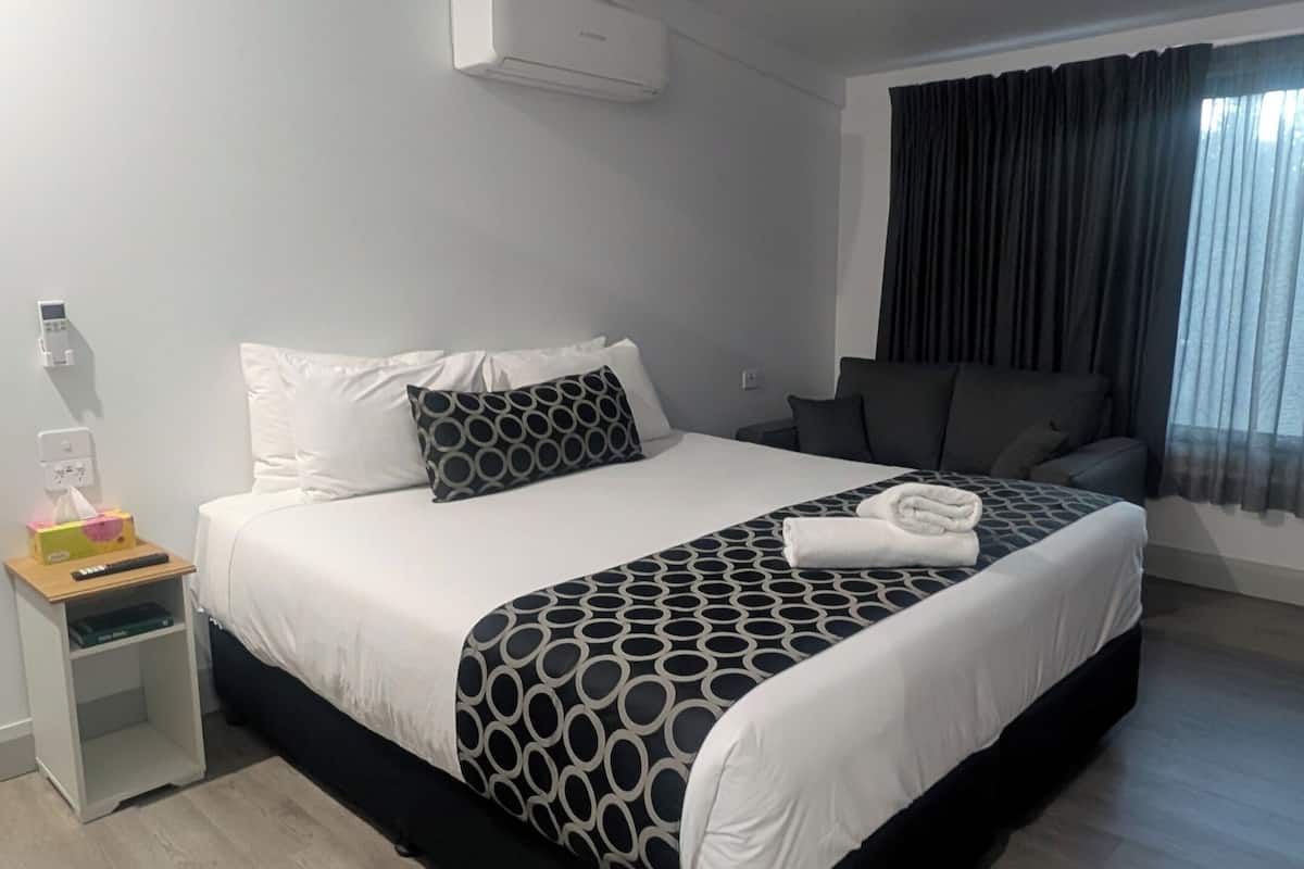 A modern hotel room with a large bed, a bedside table, an air conditioning unit, and a view of the outdoors through a window.