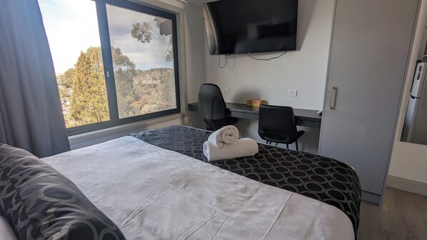 A hotel room with a bed, a desk, a chair, a TV, and a window with a view of trees. - Cooma High Country Motel (Cooma)