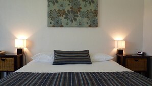 Una cama bien hecha con una colcha a rayas, dos almohadas y un tapiz floral. Hay dos mesitas de noche con lámparas y un reloj.