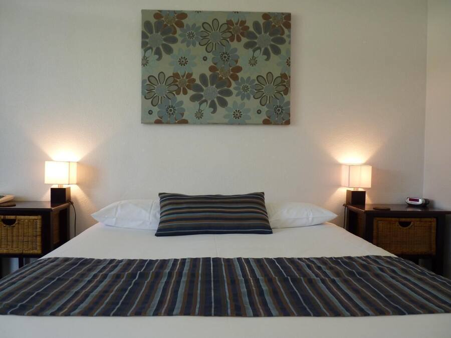 Una cama bien hecha con una colcha a rayas, dos almohadas y un tapiz floral. Hay dos mesitas de noche con lámparas y un reloj.