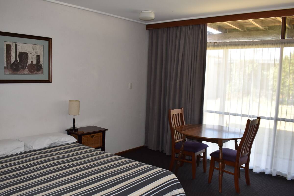 A hotel room with a bed, a table with chairs, a lamp, a framed picture on the wall, and a window with curtains.