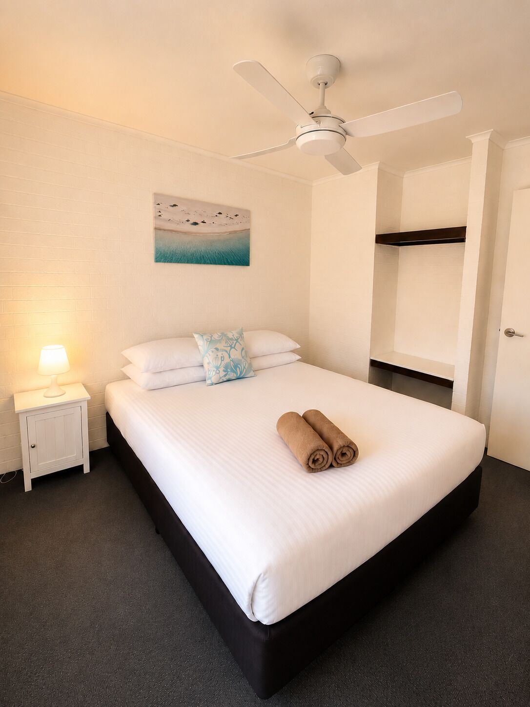 Una habitación de hotel con una cama, un ventilador de techo, una mesita de noche con una lámpara, un cuadro en la pared y una litera en la esquina.