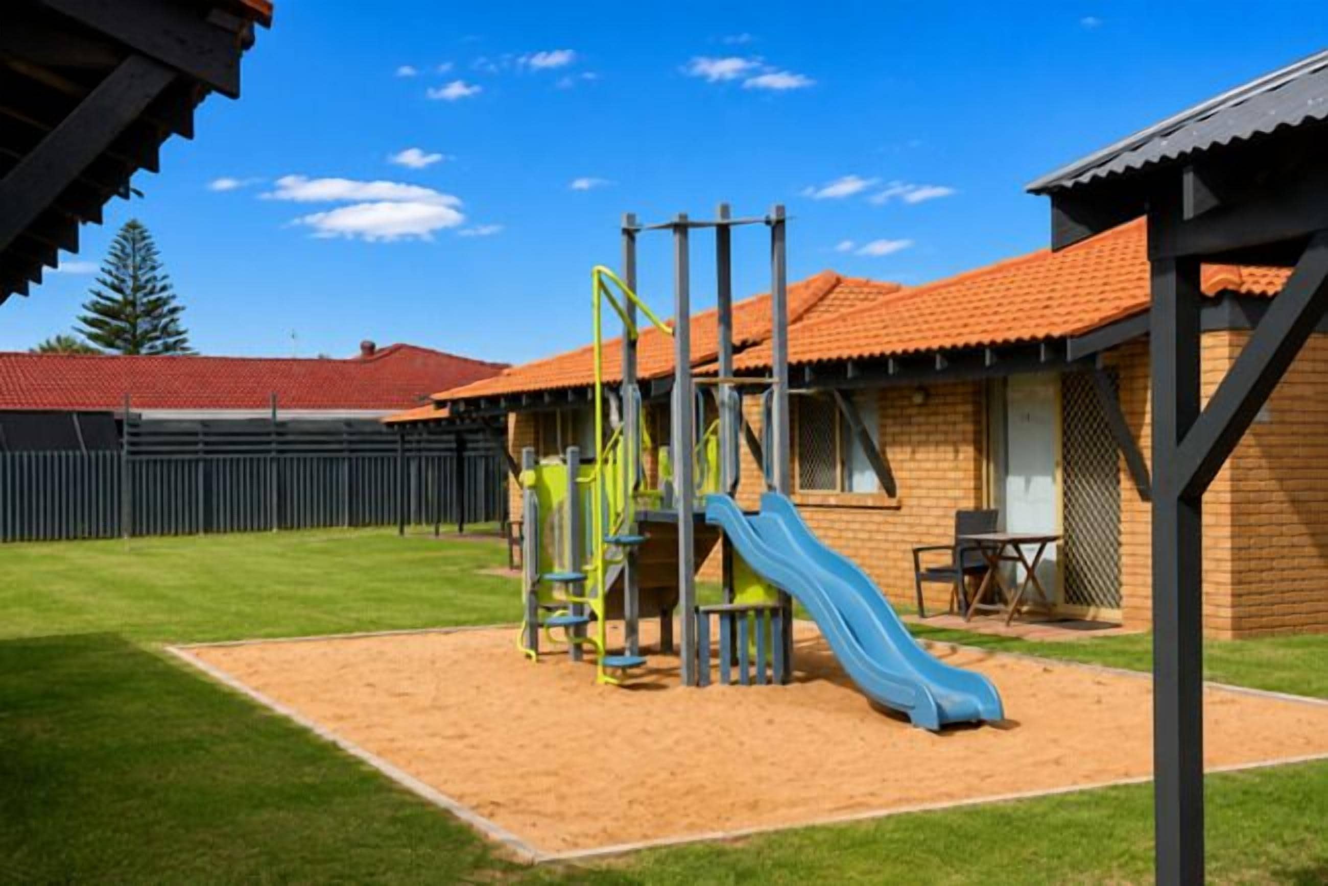 A playground with a slide and climbing structures, a fenced area, and a building with a red-tiled roof.