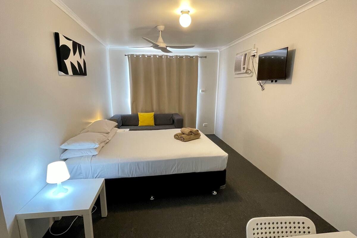 Un dormitorio con cama, mesita de noche, silla y televisión en la pared.