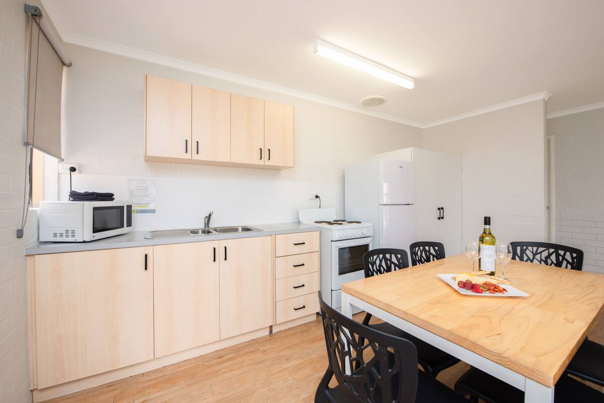 Una cocina compacta con armarios de madera clara, un microondas, una estufa y un refrigerador. Hay una mesa de comedor con sillas y una botella de vino encima.