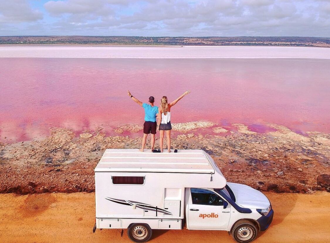 A white camper van parked on a pink beach with two people standing on top, facing a vast pink lagoon.