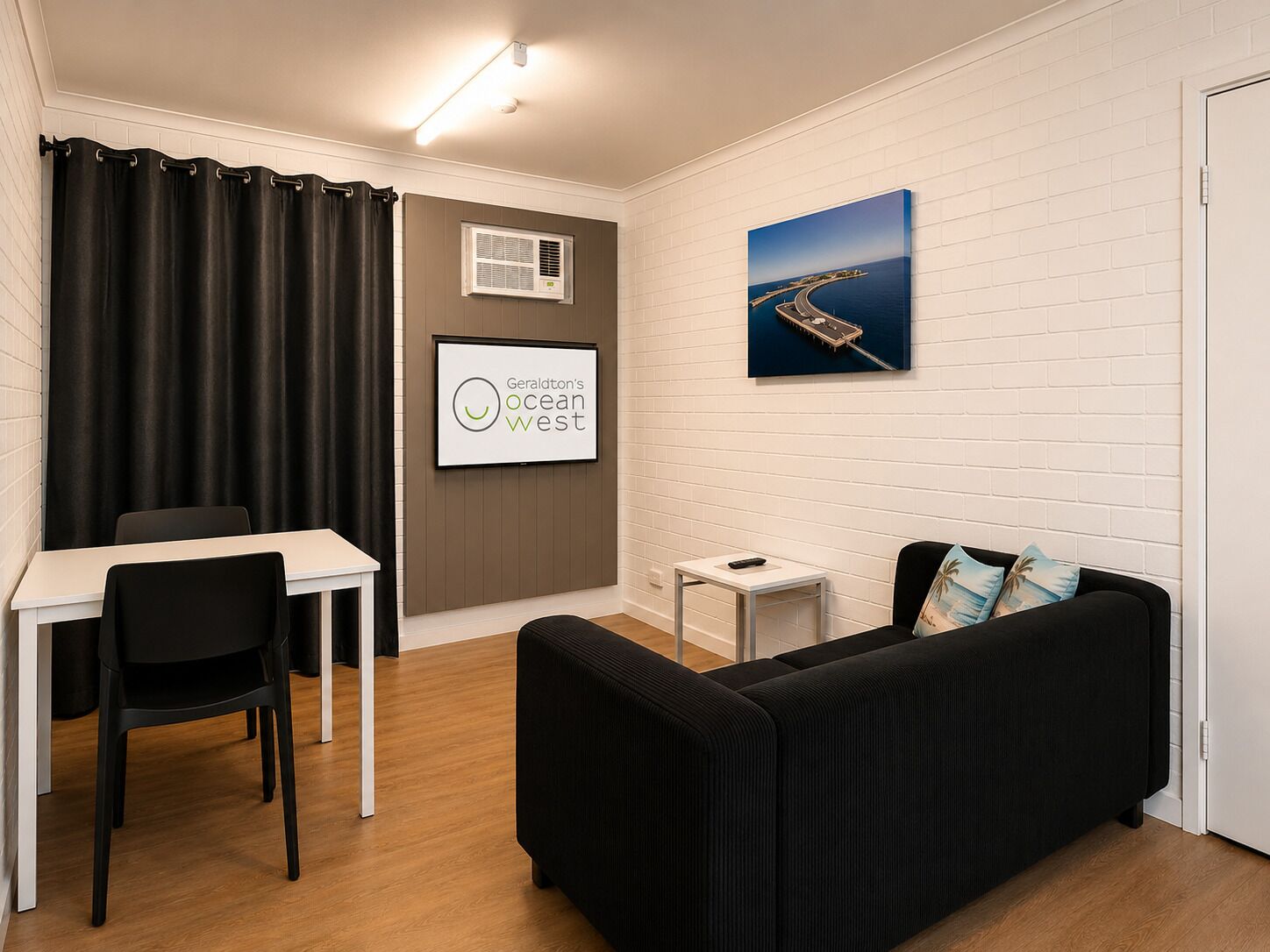 A modern room with a black sofa, a white desk, and a chair. There's a framed picture on the wall and an air conditioning unit.