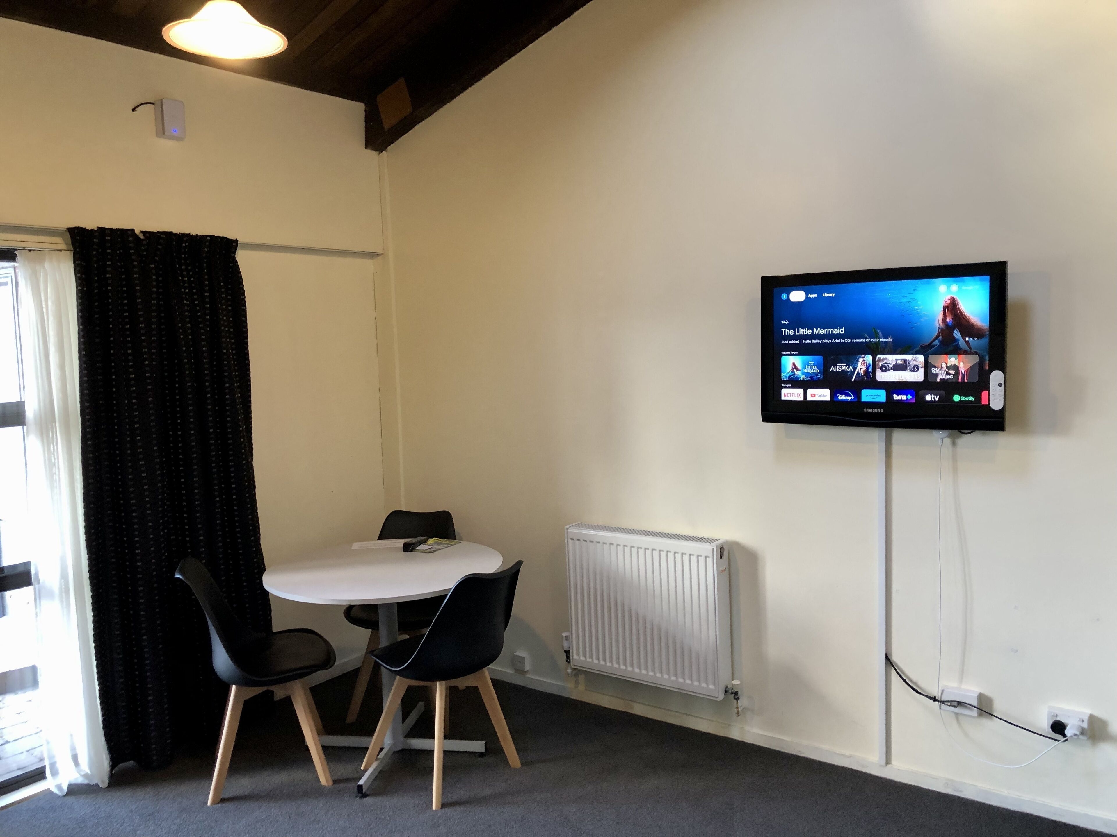 A room with a round table, two chairs, a TV mounted on the wall, a radiator, and a window with curtains.