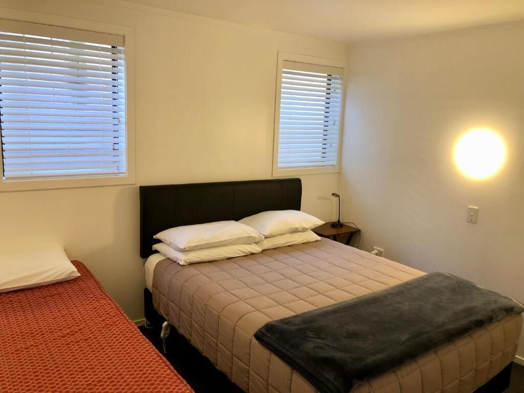 Une chambre à coucher avec un lit, deux fenêtres équipées de stores, une lampe de chevet et un luminaire fixé au mur.