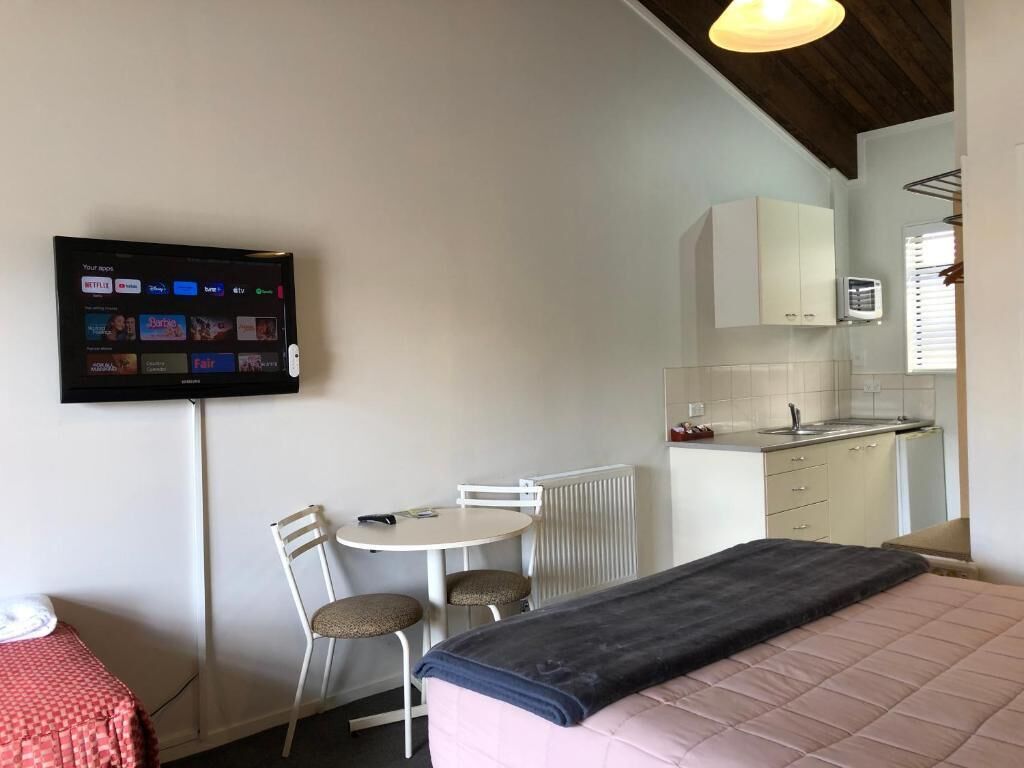 Habitación de hotel pequeña con cama, escritorio, silla, televisión montada en la pared y kitchenette.