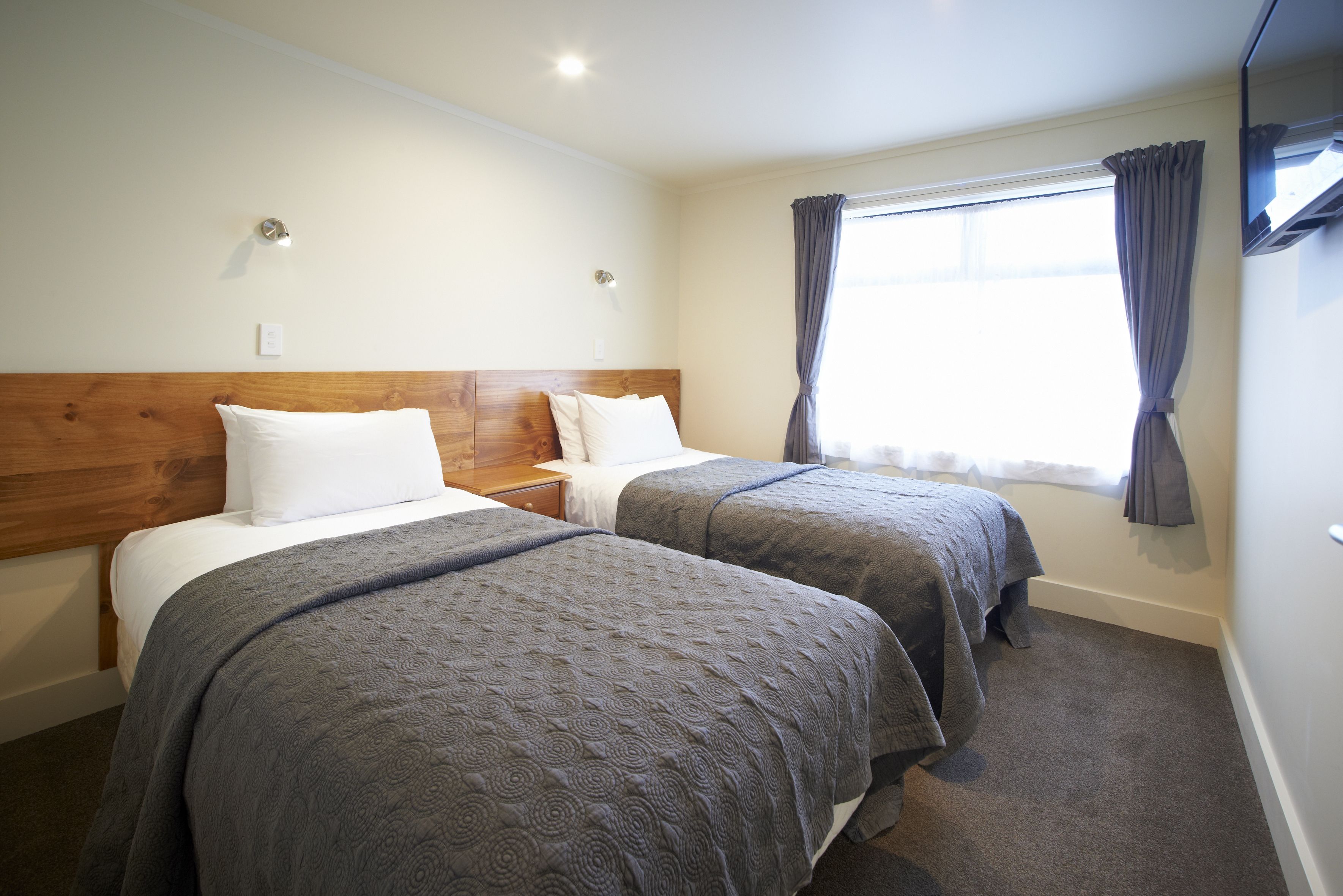 A hotel room with two beds, wooden headboards, a window with curtains, and a flat-screen TV mounted on the wall.