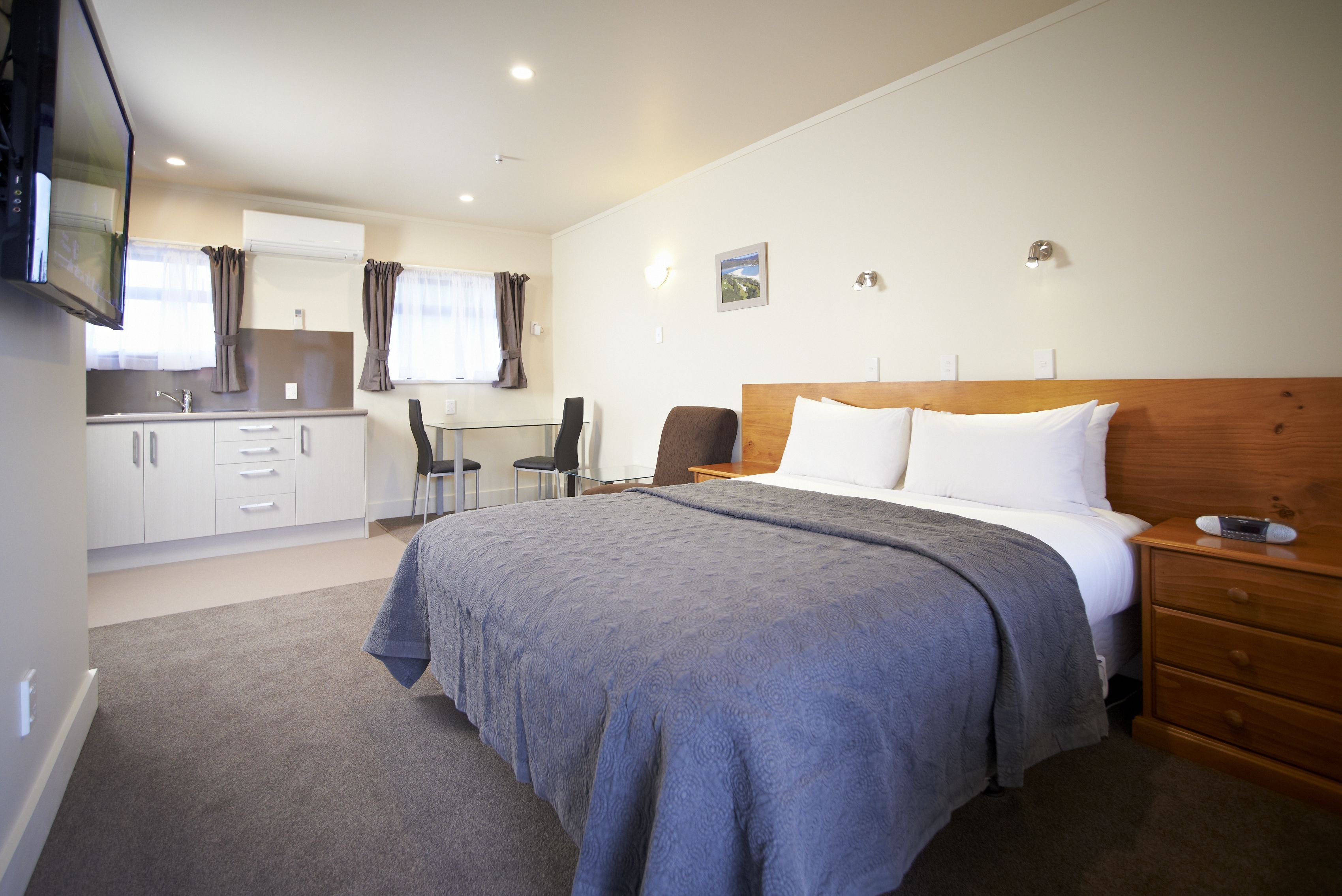 A bedroom with a large bed, a wooden headboard, a bedside table, and a nightstand. There is a small desk with a chair and a window with curtains.